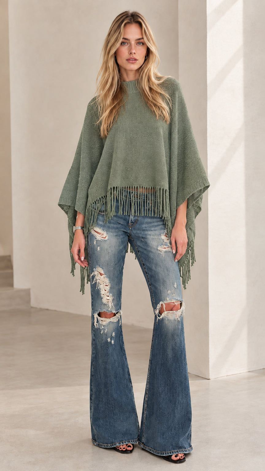 Model wearing sage green knit poncho top with fringe hem and flared denim jeans